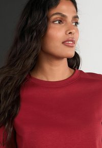 Piros pamut póló kerek nyakkivágással, sima textúrával és diszkrét varrási részletekkel. A modellen hosszú, sötét haj van, és semleges háttér látható.