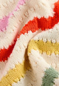 Couverture tricotée en crème, ornée de rayures en zigzag colorées en rose, orange, jaune et vert. La texture est douce et confortable.
