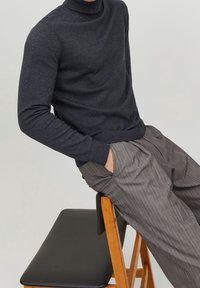 Personne portant un pull à col roulé gris foncé et un pantalon gris à fines rayures, assise de manière décontractée sur une chaise en bois avec un siège noir.