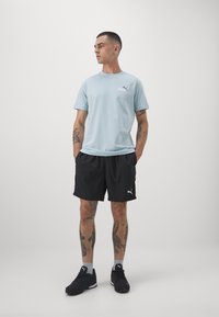 Camiseta de algodón azul claro, pantalones cortos negros y zapatillas negras con acentos blancos; presenta tatuajes visibles en los brazos. Logo de la marca en la ropa.