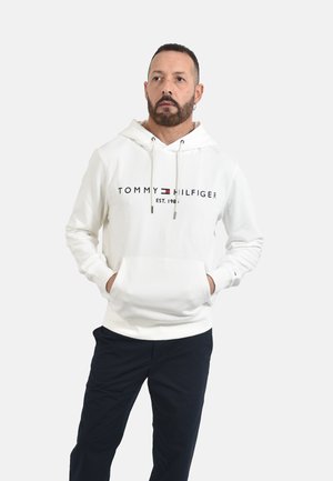 Uomo con barba che indossa una felpa bianca Tommy Hilfiger con le mani nella tasca anteriore, in piedi davanti a uno sfondo uniforme.