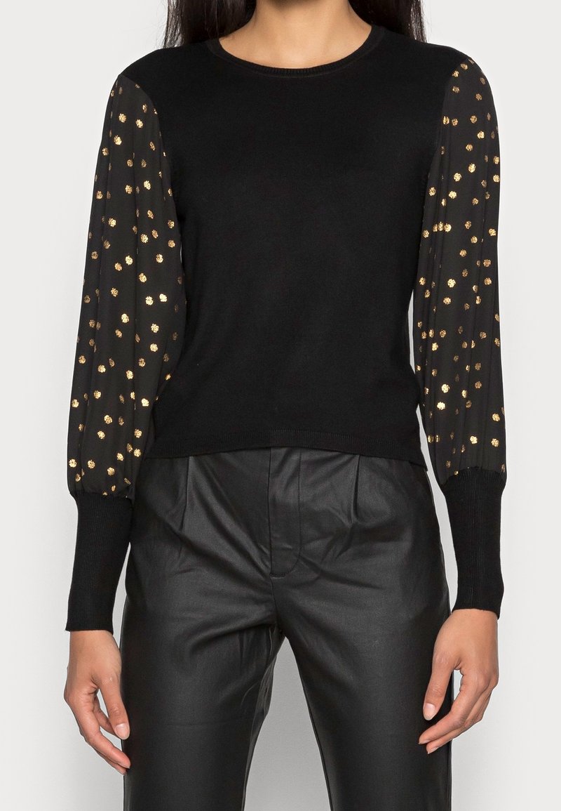 Femme portant un pull en tricot noir avec des manches transparentes noires ornées de pois dorés, assorti à un pantalon en cuir noir taille haute.