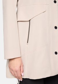 Beżowa kurtka z przednią kieszenią zapinaną na zamek, zapięciami na guziki oraz o linii A. Tkanina jest gładka z widocznymi detalami przeszyć.