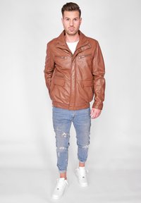 Brun läderjacka med hög krage, flera fickor med dragkedja, i kombination med slitna ljusblå jeans och vita sneakers.