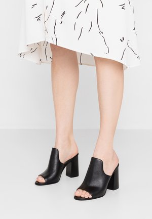 Kobieta nosząca czarne skórzane klapki na obcasach w kształcie bloku, zestawione z białą spódnicą z czarnymi abstrakcyjnymi liniami.