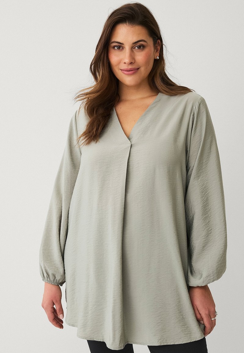Lichtgroene, lange tuniek met een V-hals en ballonsleeves. Glad, textuurstof met een A-lijn vorm en zijsplitten.