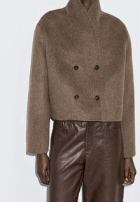 Person wearing a cropped, brown fuzzy double-breasted jacket with four black buttons and high-waisted dark brown leather pants.