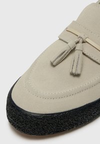 Beige suede shoe featuring a rounded toe, textured black rubber sole, and decorative tassels with a gold accent.
