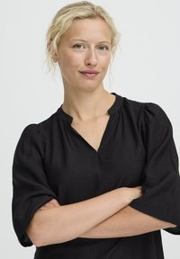Juoda bluzonė su V formos iškirpte ir trumpomis, pūstomis rankovėmis. Pagaminta iš tekstūruoto audinio, pasižymi laisvu siluetu ir gathered detalėmis ant pečių.