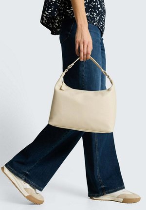 Person trägt dunkelblaue Jeans mit weitem Bein und weiße Turnschuhe, hält eine beige Handtasche am Griff und macht einen Schritt vorwärts vor einem einfarbigen Hintergrund.