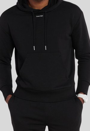 Homme portant un sweat à capuche noir Calvin Klein avec poche kangourou et pantalon noir, montre blanche au poignet gauche, mains dans les poches.