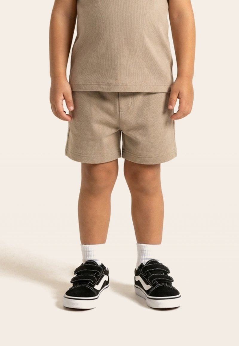 Enfant portant un short beige et un t-shirt assorti, des chaussettes blanches et des baskets noires avec semelles et détails blancs.