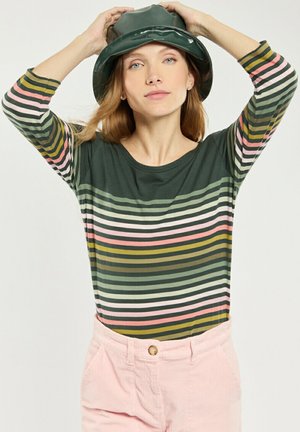 Vrouw met een groen gestreept shirt met lange mouwen en roze broek die een donkergroene bucket hat aanpast tegen een effen achtergrond.