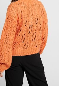 Pomarańczowy dzianinowy sweter o luźnym kroju, z teksturowanym wzorem z otwartych ściegów i ściągaczem na dole. Rękawy są szerokie i masywne.