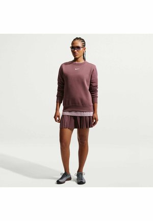 Kvinde iført en lilla Nike sweatshirt, plisseret nederdel, solbriller og grå sneakers, stående mod en ensfarvet hvid baggrund.