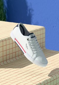 Tenis albi din pânză cu accent bleumarin, având o talpă din cauciuc, design cu sireturi și o bandă roșie pe lateral cu detalii de brand.