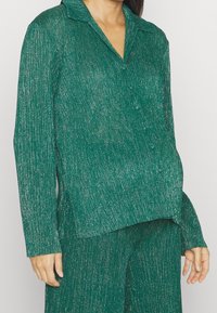 Grön metallisk långärmad skjorta med krage, med en strukturerad, skimrande tyg och en avslappnad passform. Knappdetaljer synliga.