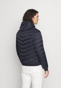 Marineblaue Steppjacke mit Kapuze, ausgestattet mit einem Zickzackmuster, elastischen Bündchen und einem taillierten Saum, getragen über weißen Hosen.