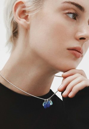 Collar de cadena de bola plateada con gotas de cristal azul y verde azulado, con una textura lisa y brillante. Lleva puesto sobre una blusa negra.