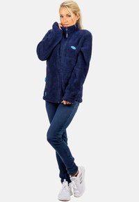 Marineblauwe fleecejack met hoge kraag, rits en zijzakken; zachte textuur met een turquoise logoaccent op de borst.
