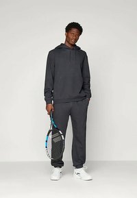 Tmavošedá mikina a zodpovedajúce teplákové nohavice vyrobené z mäkkého materiálu. Osoba drží tenisovú raketu s modrými akcentmi a má na sebe biele tenisky.