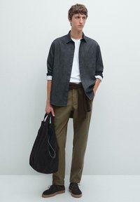 Chemise en chambray foncé, t-shirt blanc, pantalon vert olive, chaussures en daim marron et un sac de voyage marron foncé. Superposition décontractée avec une ceinture texturée.