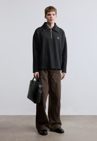 Pull-over noir à manches longues avec un col zippé, associé à un pantalon ample marron. Le modèle porte un sac en cuir noir et des chaussures noires.