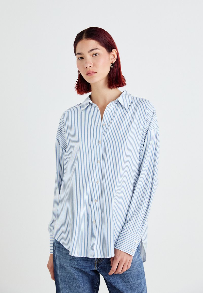 Camisa de botones azul claro con rayas verticales blancas, de manga larga, con cuello y un corte relajado. Combinada con jeans de mezclilla azul.