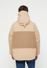 Veste beige avec un col haut, présentant un design bicolore avec une rayure marron plus foncée. Le matériau semble texturé, avec des poches fonctionnelles.