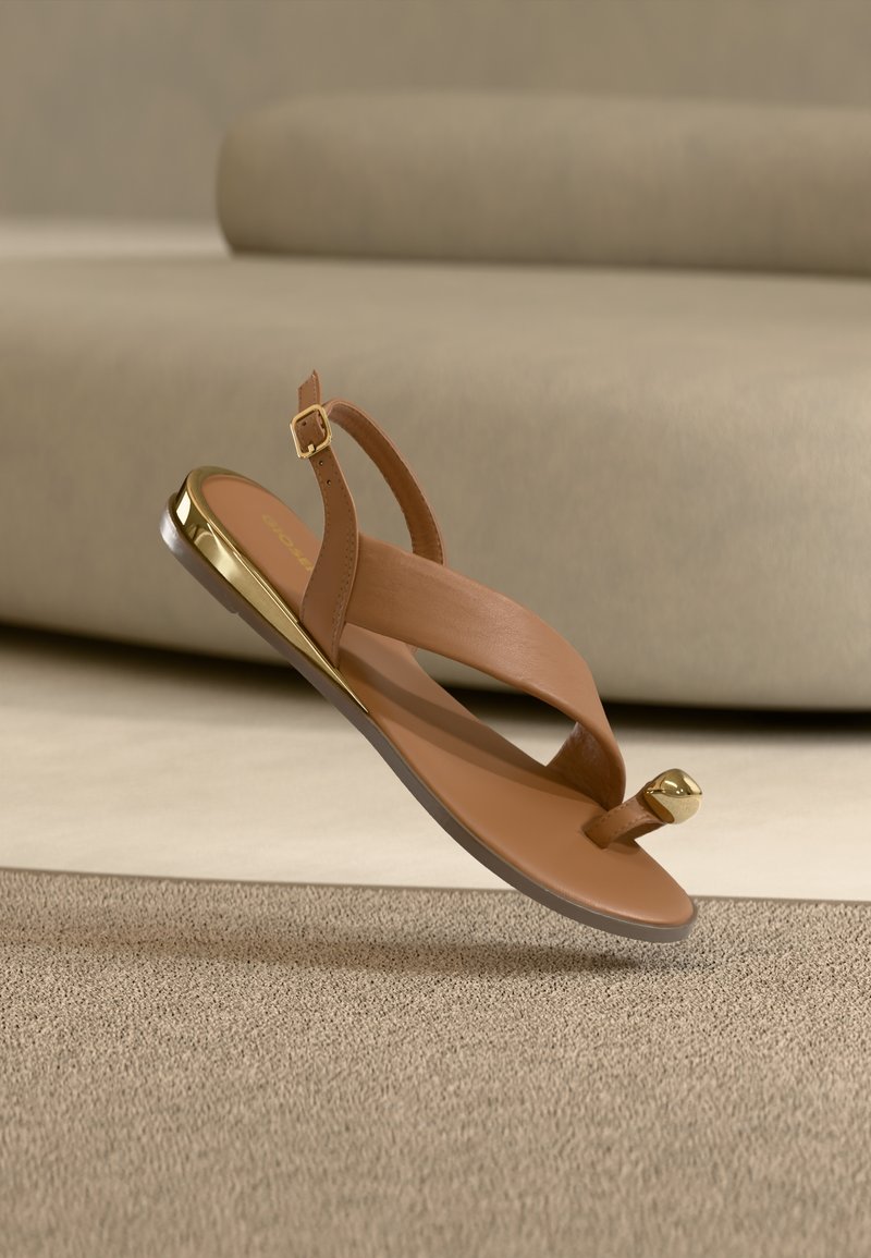 Καφετί δερμάτινο σανδάλι με χρυσή αγκράφα, δάχτυλο περασμένο στο λουράκι και μεταλλική χρυσή λεπτομέρεια στη φτέρνα πάνω σε μπεζ χαλί, με μπεζ καναπέ στο φόντο.