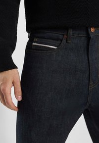 Jeans en denim bleu foncé avec une coupe droite, présentant un design classique à cinq poches, des coutures contrastées et un petit accent argenté dans la poche avant.