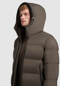 Chaqueta de plumón marrón con una gran capucha, que presenta secciones acolchadas, un cierre de cremallera y bolsillos laterales con acentos negros. Textura suave.