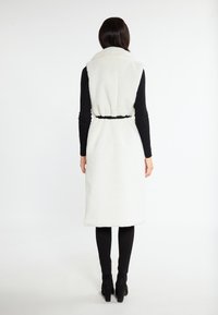 Lang, wit, fuzzy vest met een ceintuur in de taille, met een hoge kraag en een relaxte pasvorm, gecombineerd met zwarte leggings en laarzen.