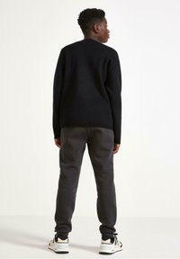 Sort strikket sweater med rund halsudskæring, parret med grå sweatpants og sort fodtøj med lyse accenter. Bageste udsigt.