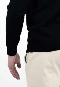 Zwarte gebreide trui met geribde manchetten en zoom, gecombineerd met lichtgekleurde broek. Hand gepositioneerd nabij de opening van de zak.
