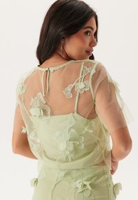 Durchsichtige mintgrüne Bluse mit floralen Stickereien und dreidimensionalen Akzenten, kurzen Ärmeln und einer Knopfverschluss auf der Rückseite.