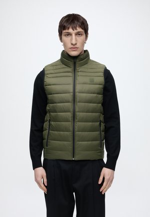 Homme portant un gilet matelassé vert olive par-dessus une chemise noire à manches longues, debout devant un fond blanc uni.
