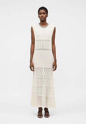 Femeie în picioare, purtând o rochie maxi albă fără mâneci, tricotată, cu modele în formă de inimă și sandale negre cu vârf deschis, pe fundal gri simplu.