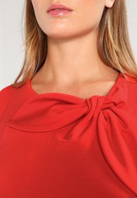 Czerwona koszula z detalem w postaci kokardy przy dekolcie, wykonana z gładkiego materiału z miękkim drapowaniem wokół ramion i klatki piersiowej.