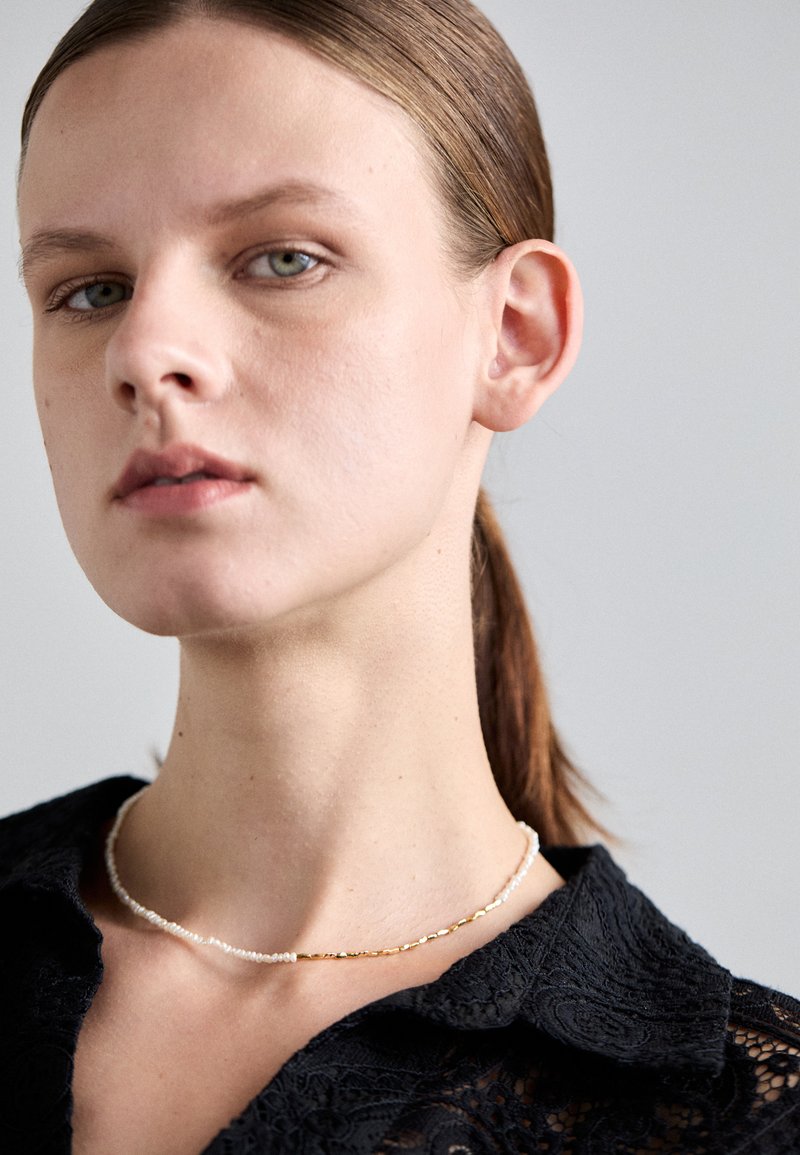 Nakit iz biserov in zlata, ki vsebuje bele bisere izmenično z zlatimi akcenti, prikazuje eleganten dizajn na črnem teksturiranem ovratniku.