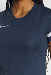 Laivastonsininen urheilupaita lyhyillä hihoilla, pyöreällä kaula-aukolla ja valkoisella logovuodolla vasemmassa olkapäässä. Sileä kankaan pinta.