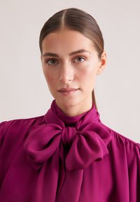 Blusa viola con un grande fiocco al collo, realizzata in tessuto setoso con una consistenza liscia, elegantemente drappeggiata con maniche a sbuffo.