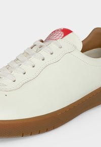 Sneakers en cuir blanc avec lacets blancs, semelle marron et patch logo rouge sur la languette avec un motif globe.