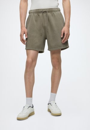 Personne portant un short vert olive à cordon, des chaussettes blanches et des baskets blanches lacées, debout sur un fond uni.