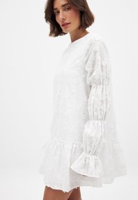 Balta siuvinėtą suknelę, papuoštą gėlių raštais, su ilgomis pūstomis rankovėmis ir išsiplėtimu apačioje. Pagaminta iš lengvos, tekstūruotos medžiagos.