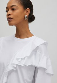 Weißes T-Shirt mit Rüschendetails an der linken Schulter, aus glattem Baumwollstoff. Model trägt einen silbernen Ohrring.