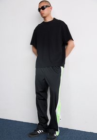 Fekete, túlságosan nagy póló, fekete sportnadrág neon zöld oldalsávokkal és fekete sportcipő. A modellen fekete napszemüveg van, minimalista dizájnnal.