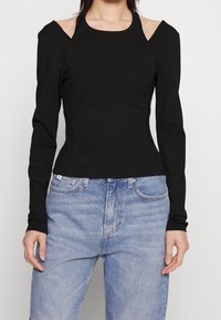 Haut noir à manches longues avec design à épaules découpées, corsage ajusté, texture douce, associé à un jean taille haute bleu clair.