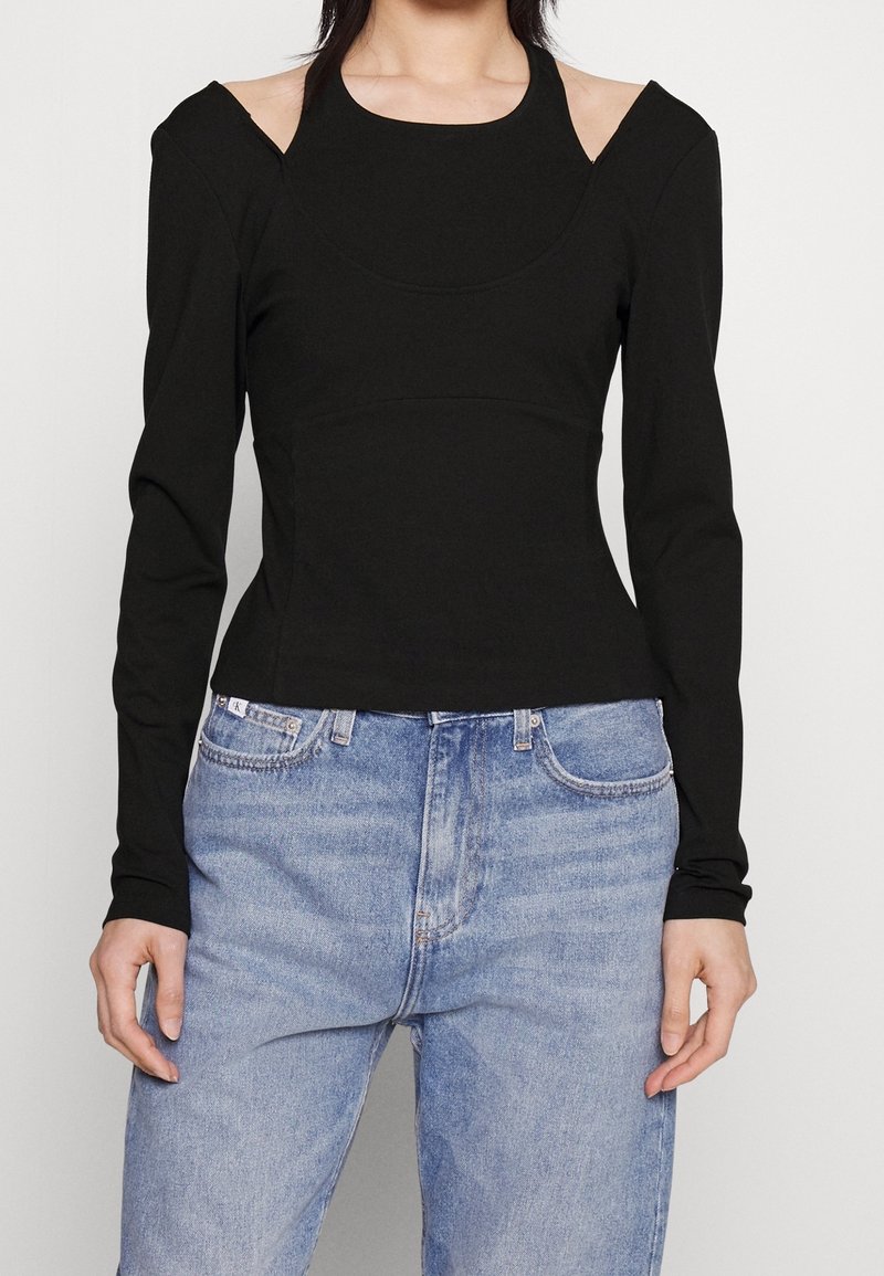 Haut noir à manches longues avec design à épaules découpées, corsage ajusté, texture douce, associé à un jean taille haute bleu clair.