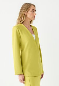 Limoengroene blazer met een V-hals, lange mouwen en gestructureerde pasvorm. Glad materiaal met minimale naden en geen zichtbare hardware of accenten.